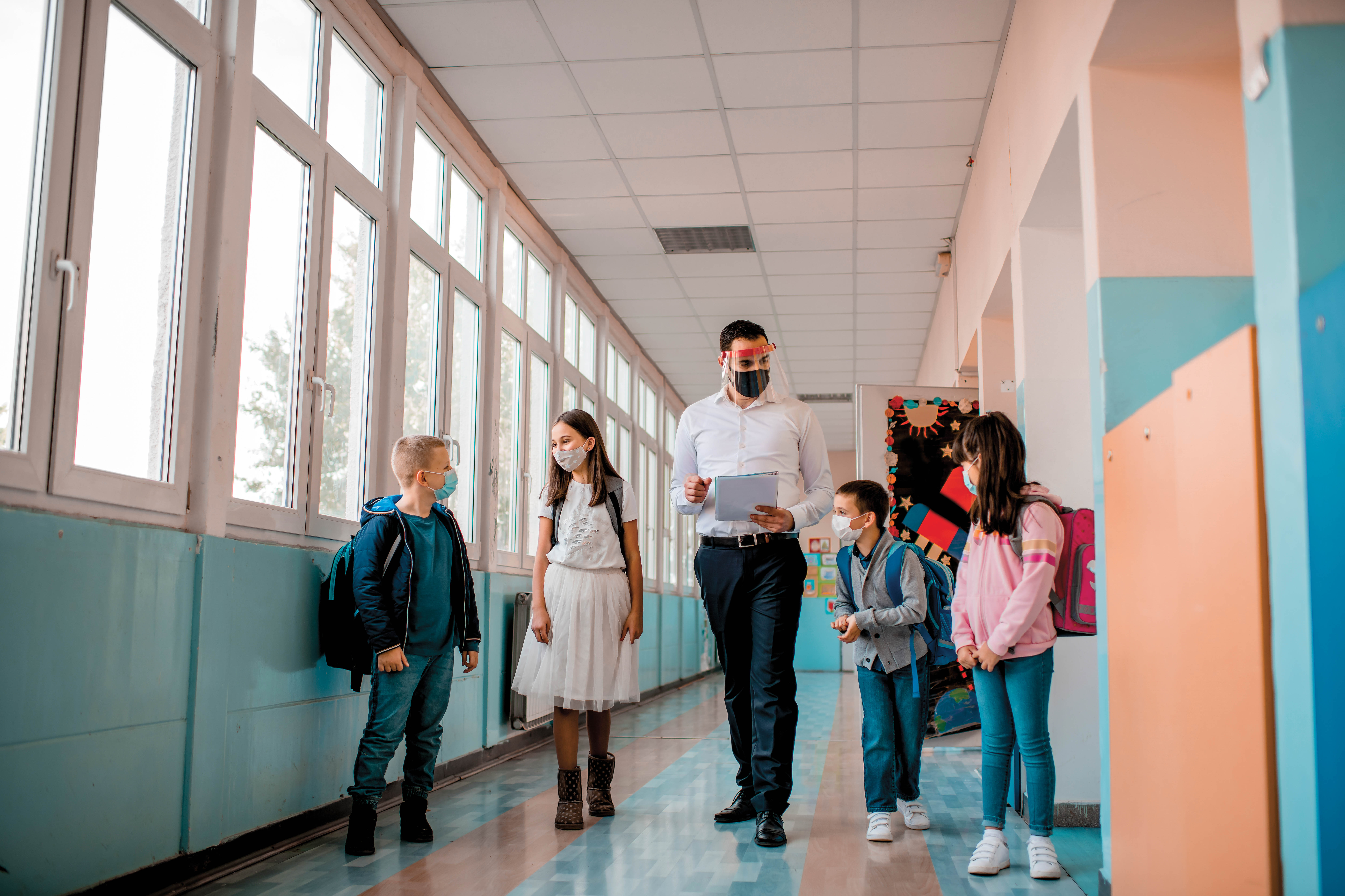 Un enseignant marchant dans un corridor. Il est entouré de quatre jeunes élèves. Ils portent tous un masque.
