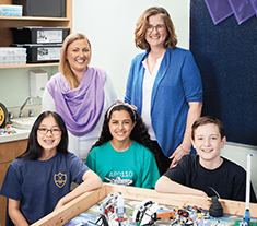 Photo of Shauna Nelson, Ontario Certified Teacher, and Sari McDowell, Ontario Certified Teacher, standing behind three students in a robotics classroom. In front of them are several robots made out of Lego.